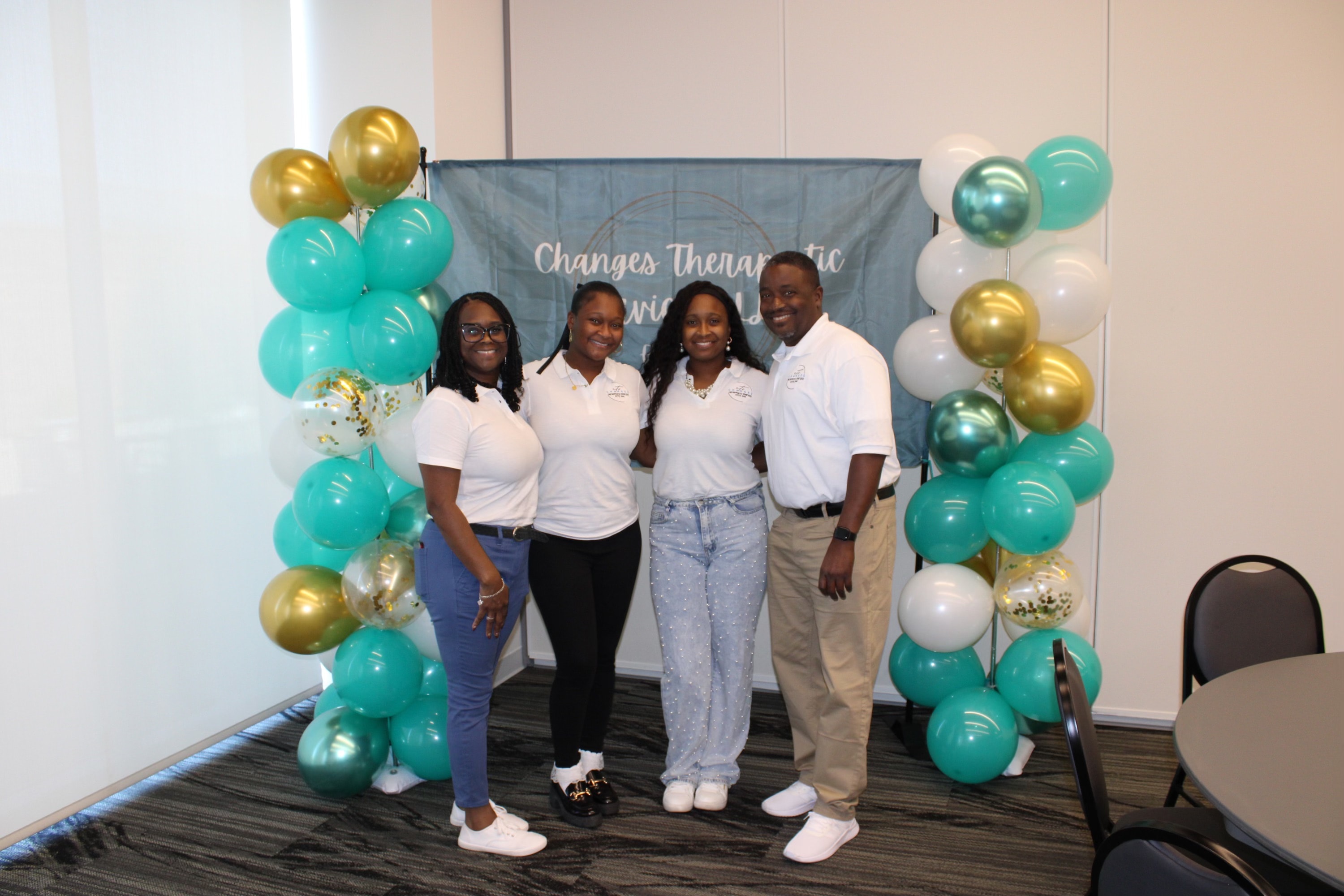 Four Changes Therapeutic Services team members posing in front of a teal, gold, and white balloon backdrop