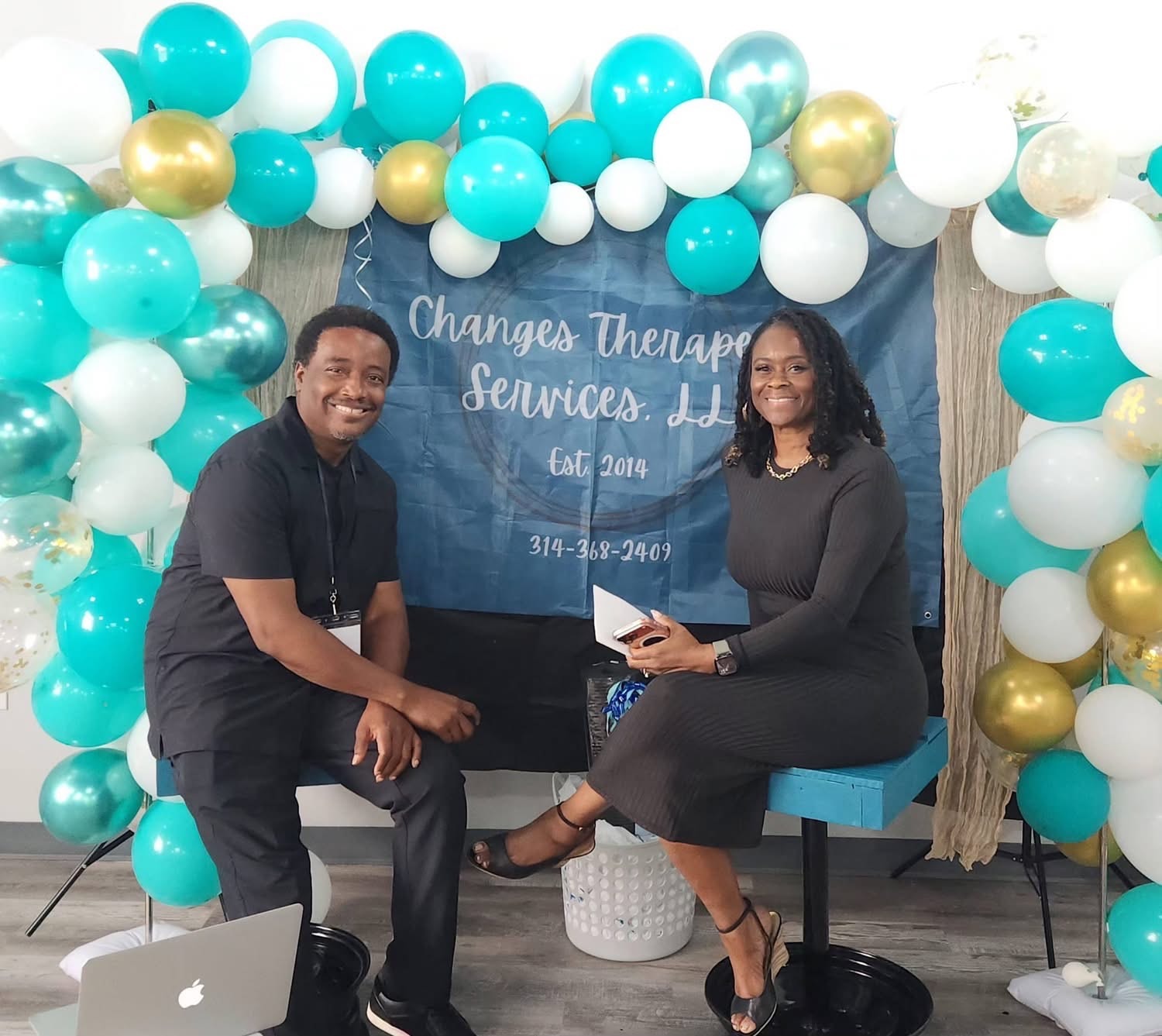 Two people seated at a Changes Therapeutic Services event display with teal, white, and gold balloons