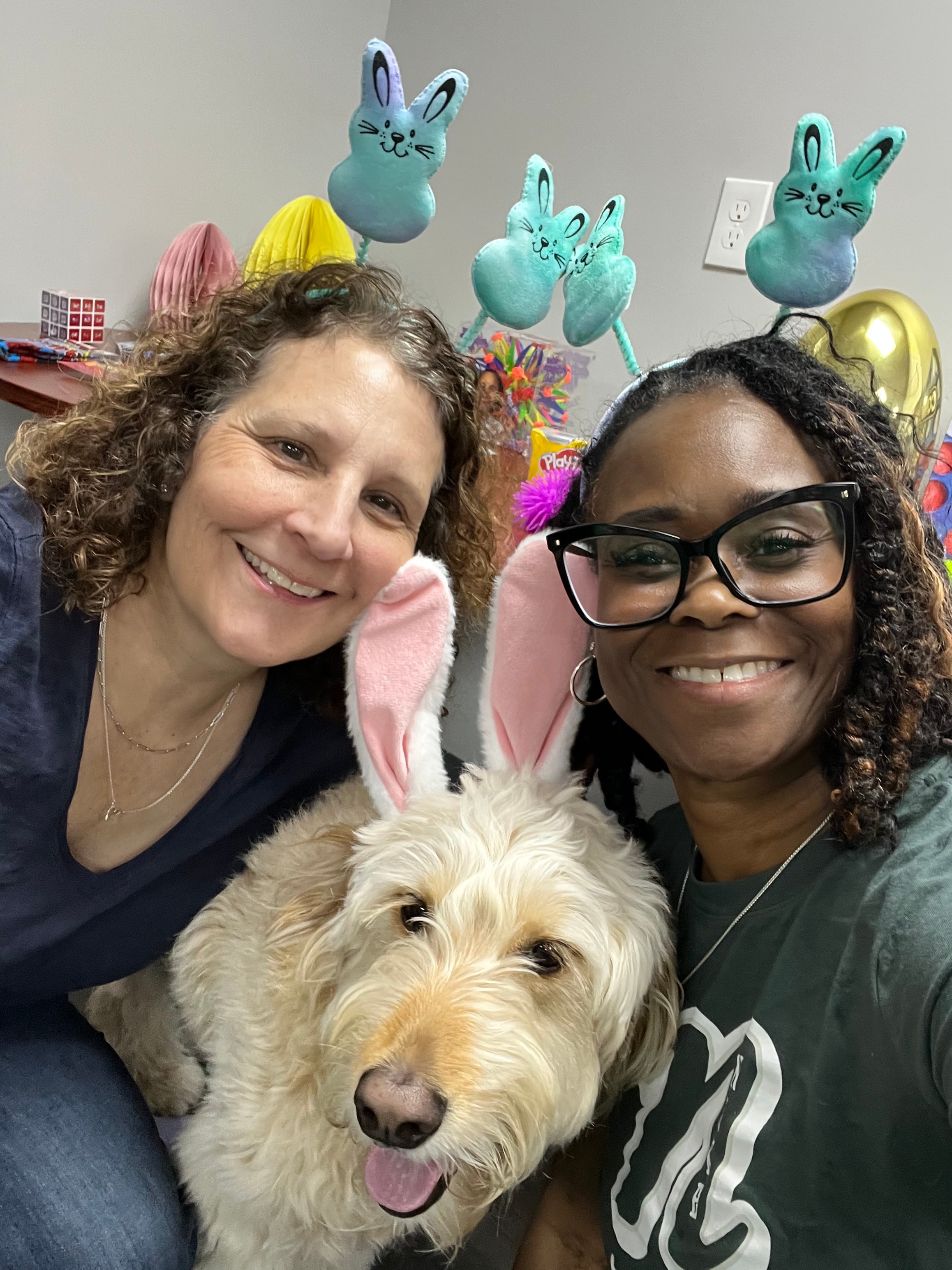 Two smiling participants and a therapy dog wearing bunny ears at an Easter event